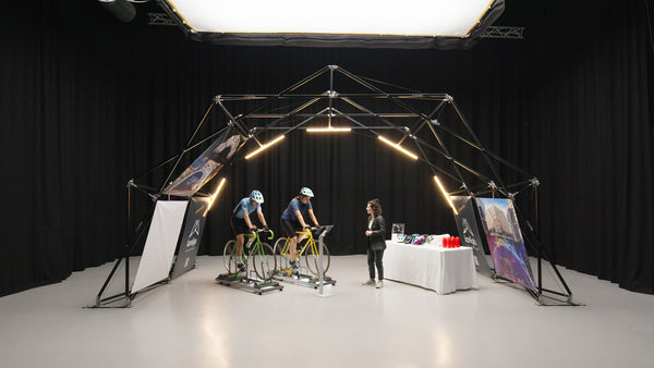 Indoor setup of Konligo event structure featuring two cyclists on stationary bikes and a display table with helmets and water bottles. Now available on MTN Shop US.